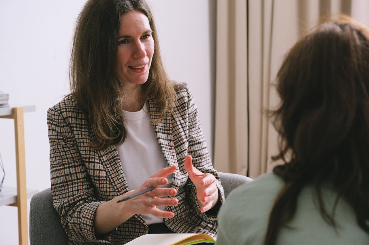 Pensive woman psychologist with brown hair in stylish clothes sitting and talking with unrecognizable female in light room