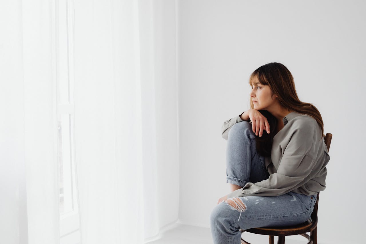 Pensive young woman in casual attire sitting on a chair indoors, contemplating.